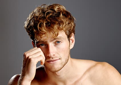 A portrait of a smiling young man with clear, healthy-looking skin against a neutral background. The image represents men's aesthetic treatments, skin rejuvenation, anti-aging services, and professional cosmetic care.
