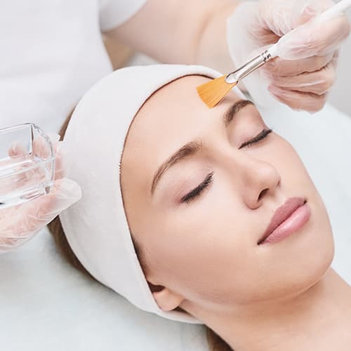A close-up of a woman receiving a professional chemical peel facial as a skincare specialist applies a treatment solution to her forehead with a brush. The image represents advanced skin rejuvenation treatments for improving skin texture, tone, acne, pigmentation, and fine lines.